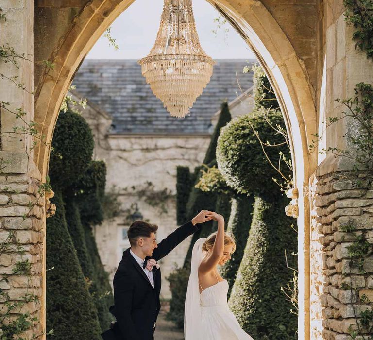 Groom in classic black grooms tuxedo spinning bride in Watters strappy sleeveless princess wedding dress and chapel length wedding veil in stone archway at Euridge Manor wedding venue with crystal chandelier above 