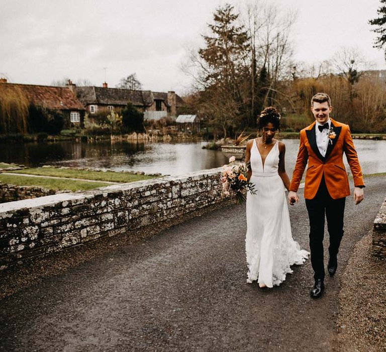 Bride in v-neck sleeveless wedding dress with 3D floral applique train walking on the grounds of Brinsop Court wedding venue with groom wearing crushed burnt orange velvet grooms blazer, black bowtie and black suit trousers 