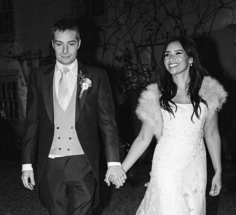 Bride in a-line tulle wedding dress with beaded lace applique with petals and a square neckline with side tulle cutouts and faux fur bridal coverup hand in hand with groom in navy grooms morning suit with grey waistcoat, off-white tie and mixed wildflower boutonniere at South Farm wedding