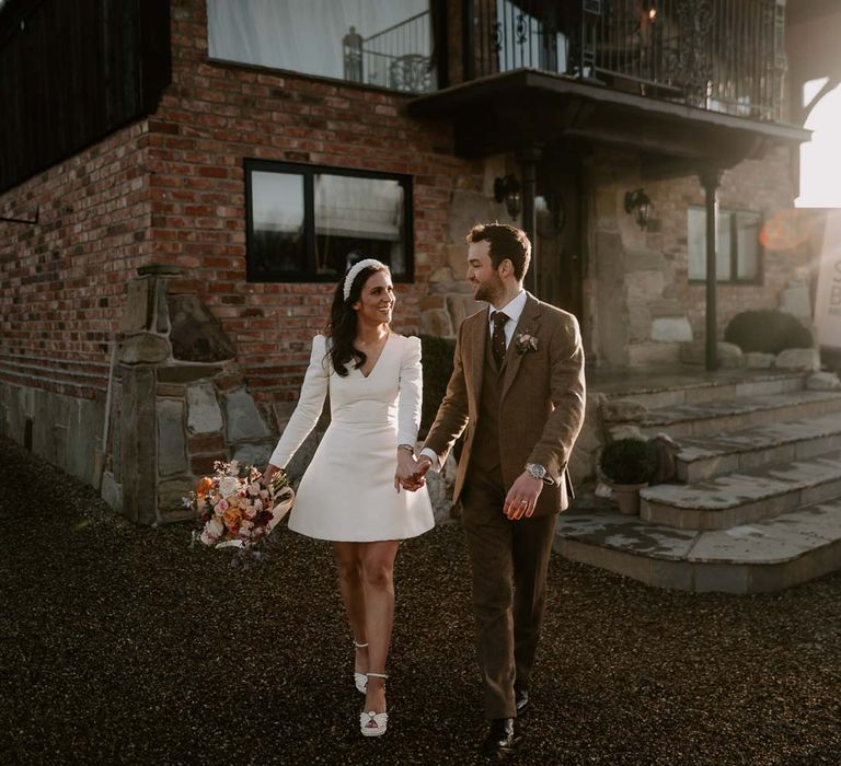 Groom in chocolate brown grooms suit walking with bride in long sleeve short wedding dress, pearl bridal headband and platform pearl wedding heels outside Willow Marsh Farm Loughborough wedding venue