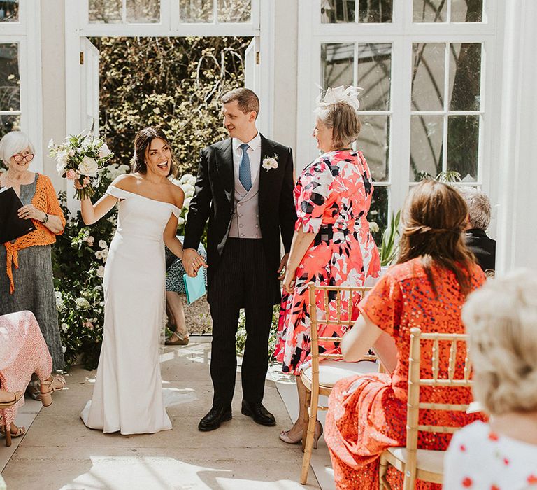 Bride in off the shoulder Halfpenny London wedding dress with white flower wedding bouquet turning with the groom to face their guests