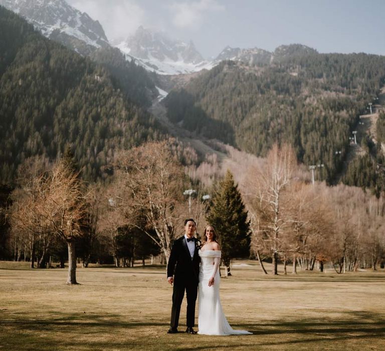 Bride in off shoulder sparkly mermaid wedding dress standing with groom in classic black tuxedo, black bowtie and white garden rose boutonniere at French destination wedding