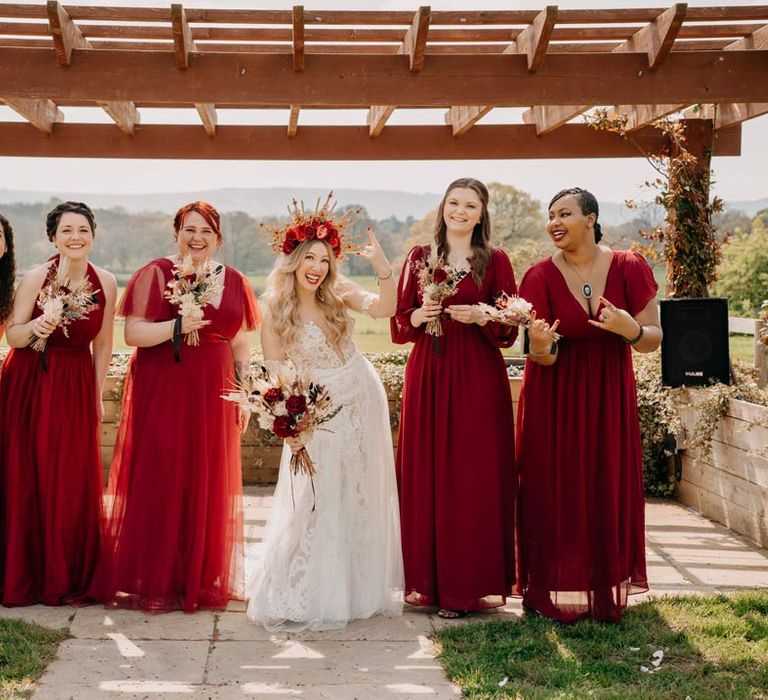 Bride in off shoulder lace wedding dress with corset style top and red rose and gold halo bridal crown holding red rose, pampas grass and dried flower bridal bouquet standing with bridesmaids in deep red mismatched bridesmaid dresses