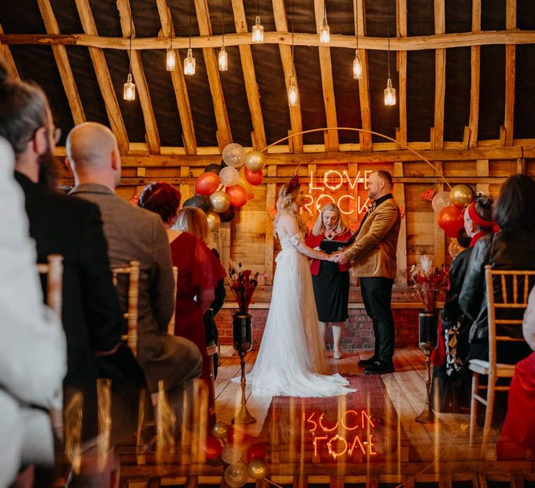 Groom in crushed velvet burnt orange grooms blazer and dried flower boutonniere and bride in off shoulder lace wedding dress with corset style top and red rose and gold halo bridal crown at the alter of Southlands Barn wedding venue reception room with hanging bottle lights, a balloon wedding arch and a large pink wedding neon light