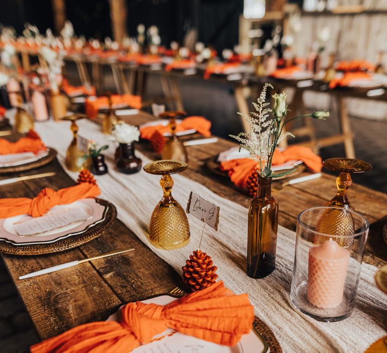 Autumn wedding tablescape with an orange napkin, pinecone wedding place name, gold charger plates and brown coloured glassware 