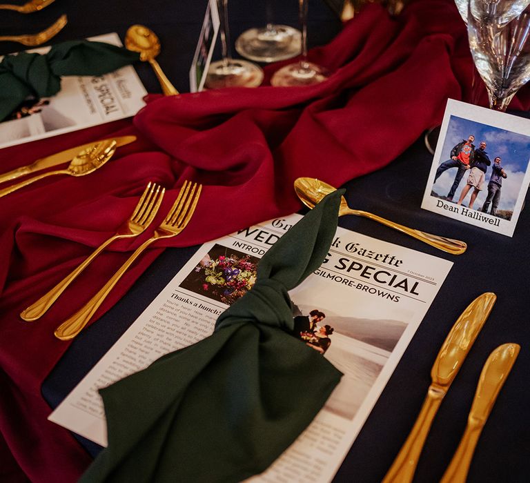 Green wedding napkin with gold cutlery and red wedding tablecloth for jewel tones alongside a wedding newspaper and Polaroid place names 