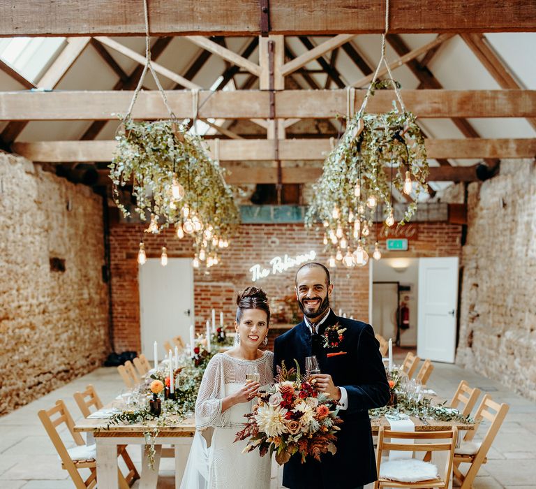 Bride & groom stand in rustic wedding venue with wooden tables and hanging lights