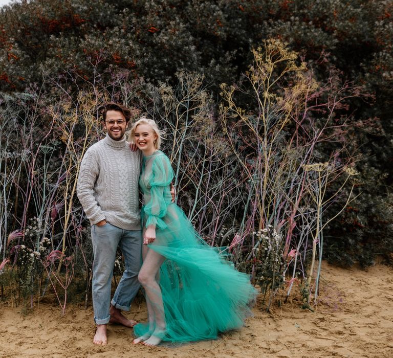 White bearded groom with glasses in a jumper and jeans on the beach with his bride in a sheer pastel wedding dress