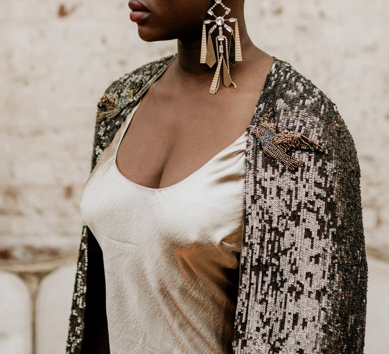 Stylish bride with alopecia in a satin wedding dress and black bridal cape with gold crown and statement earrings 