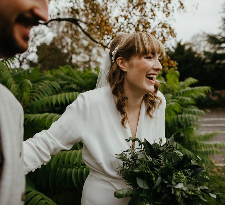 smiley bride in a wrap jumpsuit with blunt fringe and wavy hair holding a green foliage wedding bouquet