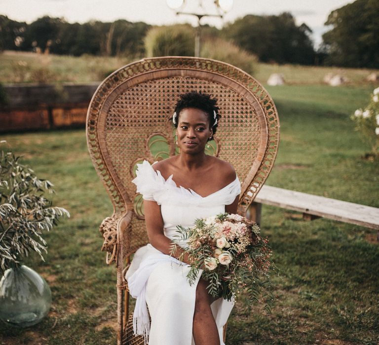 West African bride in an off the shoulder wedding dress with feather detail sitting on a wicker peacock chair 