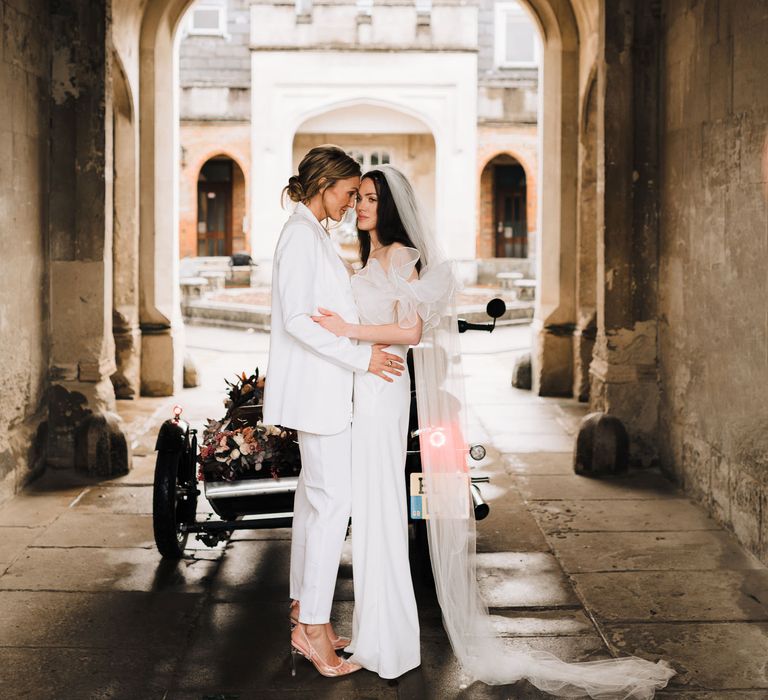 Two brides in a strapless jumpsuit and veil and trousers suit with jacket embracing at Ashridge House 