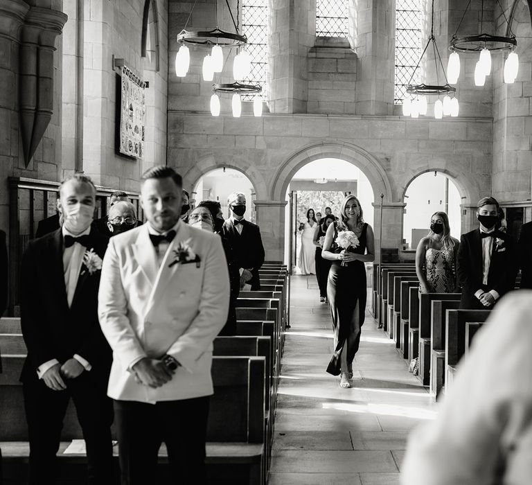 Bridesmaid in black dress holding white rose bouquet walks up the aisle towards groom in white double breasted tuxedo jacket and bow tie