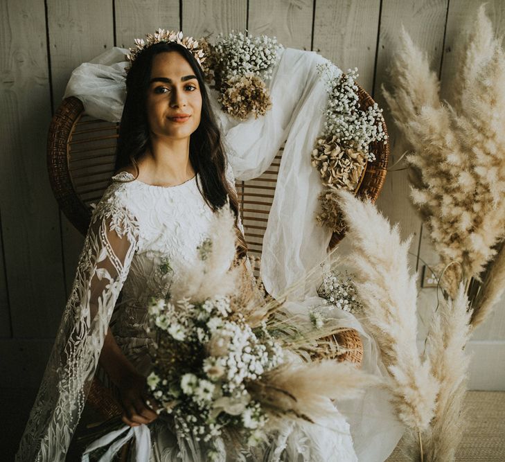 Bride in a gold leaf headdress sitting on a peacock chair surrounded by pampas grass and dried flower arrangements 