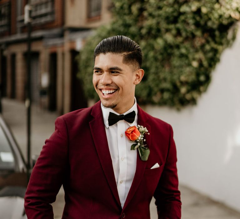 Stylish groom in a deep red tuxedo jacket and bow tie with a coral rose buttonhole