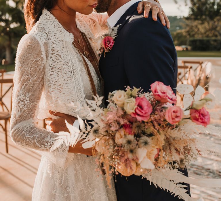 Boho bride in a lace Marylise Bridal gown and jacket embracing her husband whilst holding a pink and white wedding bouquet