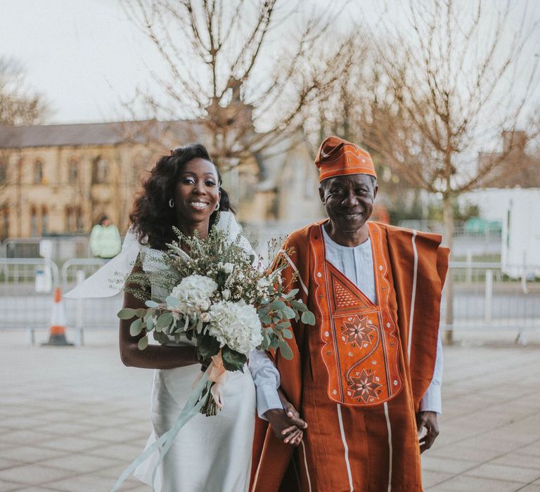 Bride in customised white Bec + Bridge wedding dress with shoulder bows, white and green wedding bouquet and curled wedding hair walks arm-in-arm with guest in traditional orange and white outfit