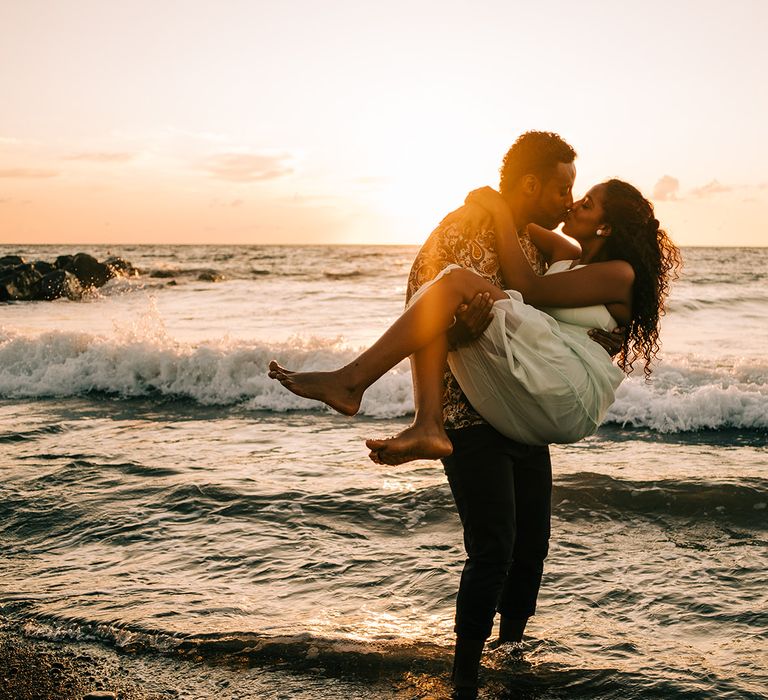 Groom carries bride along the beach as the sun sets behind them