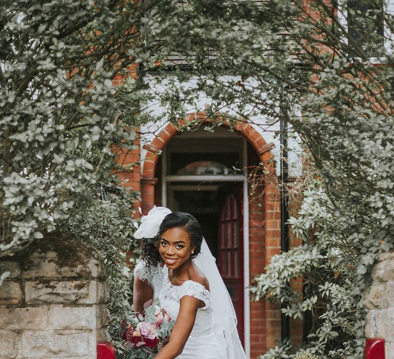 Bride in a fitted lace wedding dress and veil walking out of a red house gate