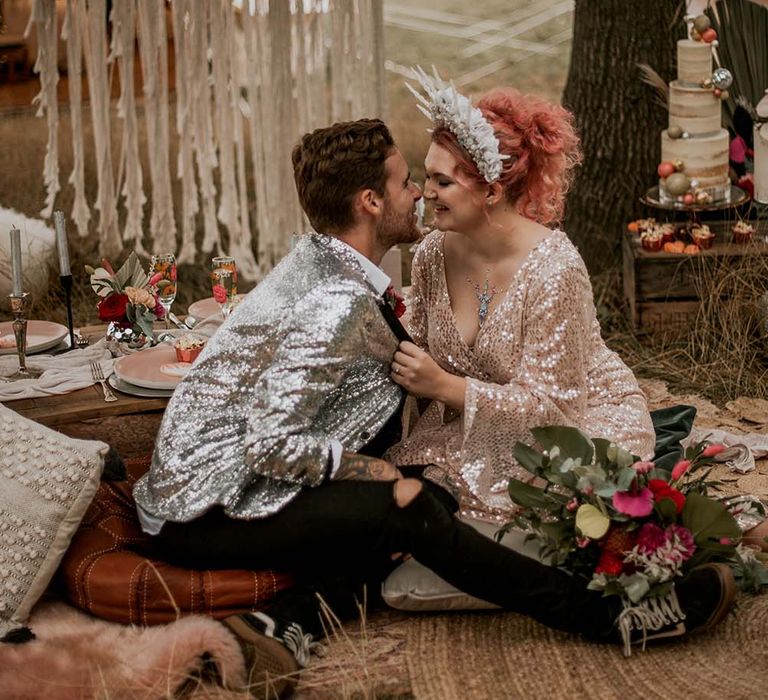 Bride with pink hair and rose gold long sleeved V neck sequin wedding dress sits holding the front of grooms silver sequin suit jacket at festival themed wedding