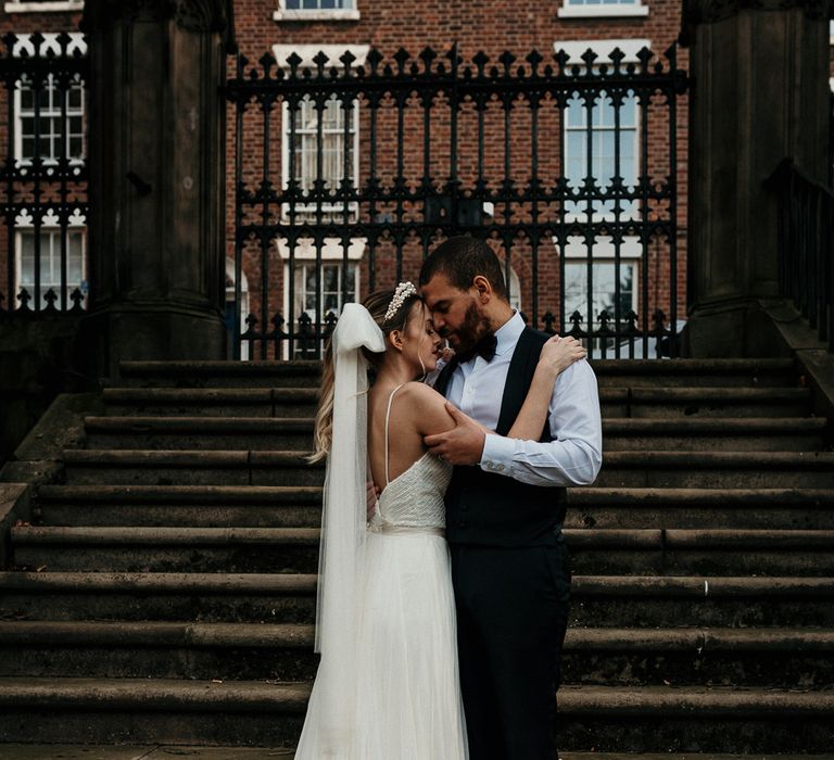 Groom in a horseshoe waistcoat and bow tie embracing his bride in a thin strap wedding dress with bow veil 