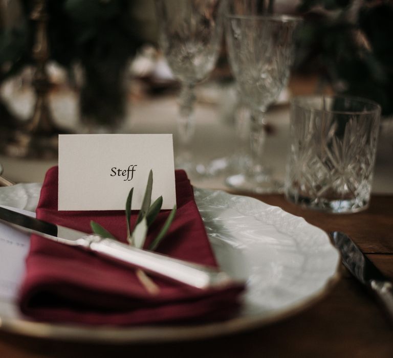 Place setting with maroon napkin, white China plate and silver cutlery, with classic white name card & sprig of rosemary 