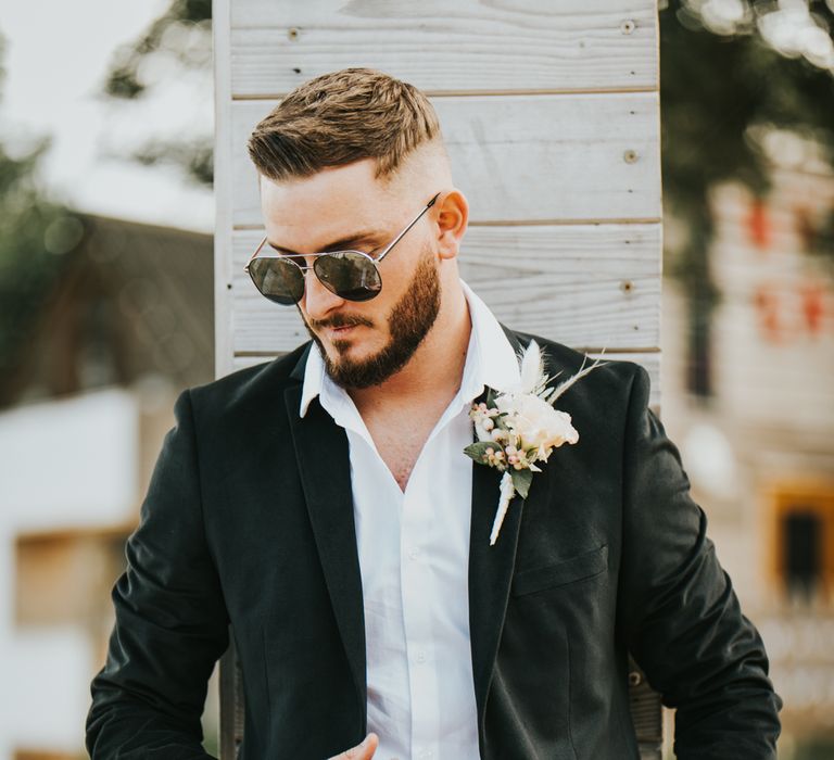 Stylish groom in a black suit, white open collar shirt and aviator sunglasses with a rose buttonhole 