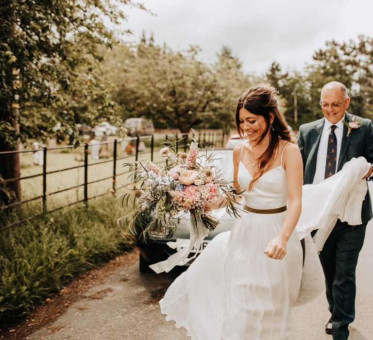 Bride with long brown curled hair and white cami wedding dress with gold ribbon waistband holding multicoloured bridal bouquet walks towards wedding whilst man in blue suit and tie holds her train