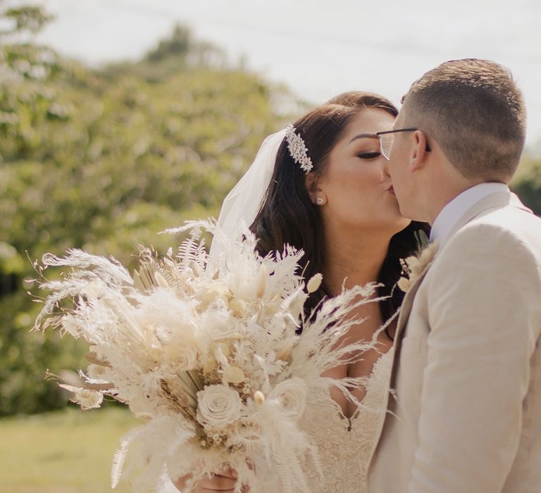 Bride with side swept wedding hairstyle holding a neutral dried flower wedding bouquet whilst kissing her groom 