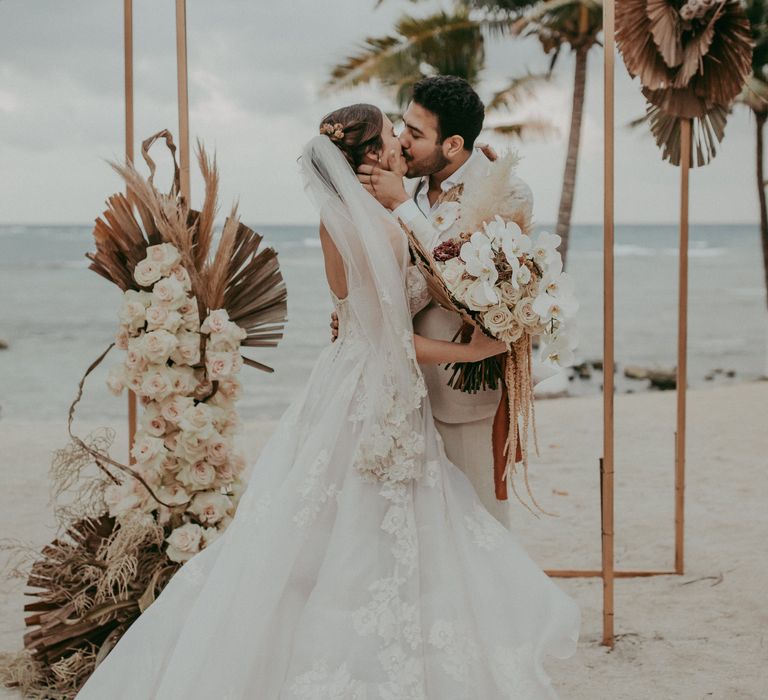 Bride & groom kiss in front of the beach next to dried florals and palm trees on their wedding day