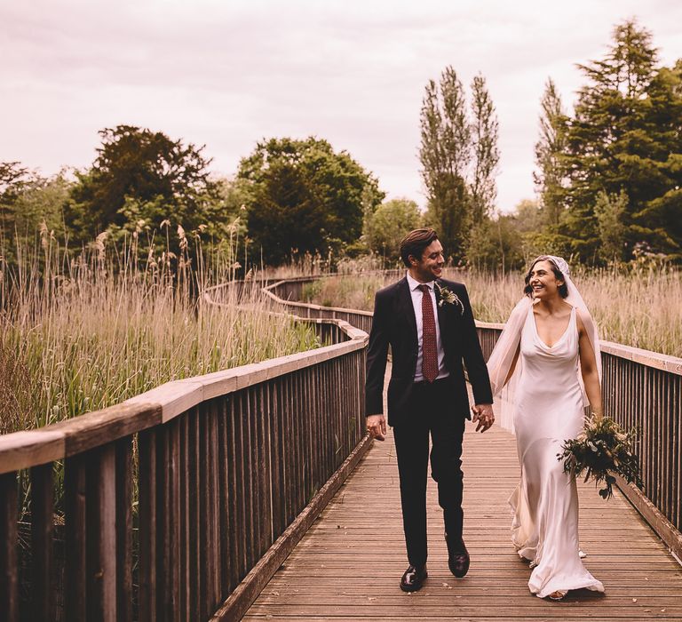 groom in a black suit with red tie holding hands with his bride in a satin wedding dress with cowl front and Juliet cap wedding veil on a bridge