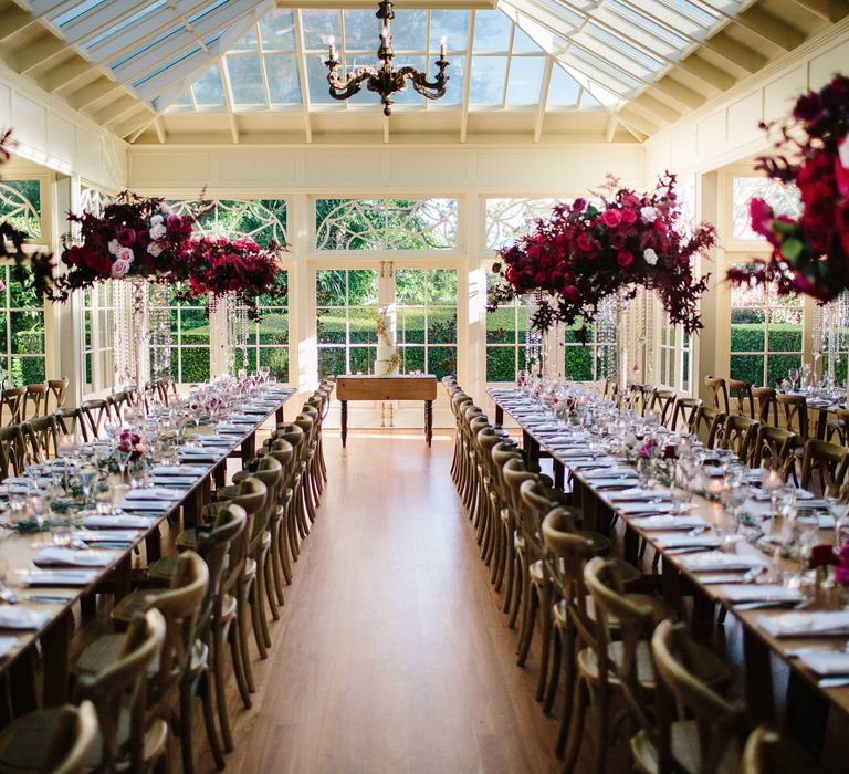 Wedding reception room lit brightly by the outside sky through large windows with bright red and pink floral decoration upon tables