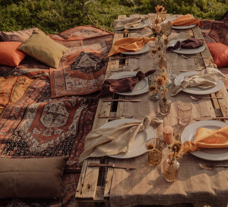 Rustic table setting on wooden palette complete with Moroccan style rugs for sitting, neutral toned fabric napkins and dried floral arrangements 