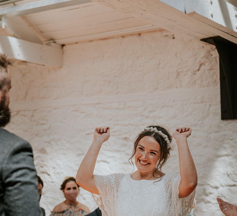 Bride lifts her hands in the air to groom as she celebrates wedding day whilst wearing bridal crown and sequin embellished wedding gown