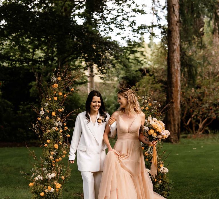 Two brides in a white trouser suit and blush wedding dress walking up the outdoor aisle with yellow vertical flower arrangement 