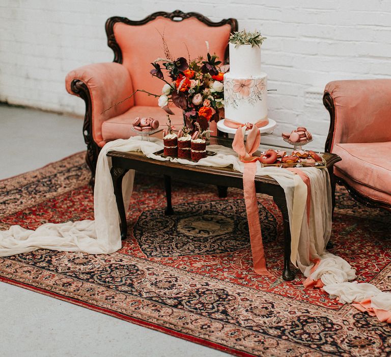 Muted pink and orange dessert table and seating area with painted wedding cake, silks, ornate chairs and wool rug 