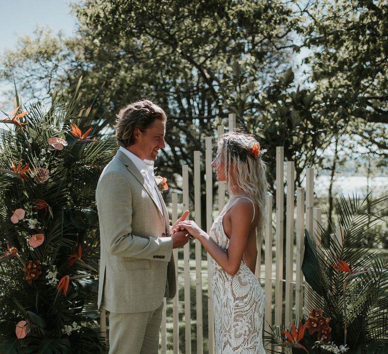 Bride in open back lace boho wedding dress with train holds hands with groom in linen suit in front of white wooden posts and tropical flower installation during outdoor wedding ceremony for birds of paradise wedding inspiration