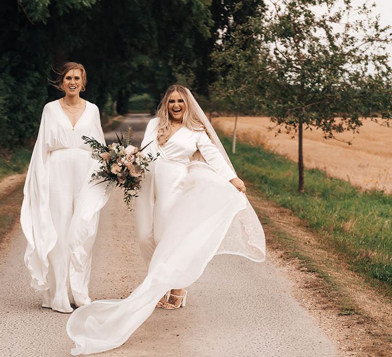 Brides walk along together as bride wears cape with jumpsuit and blonde bride wears veil