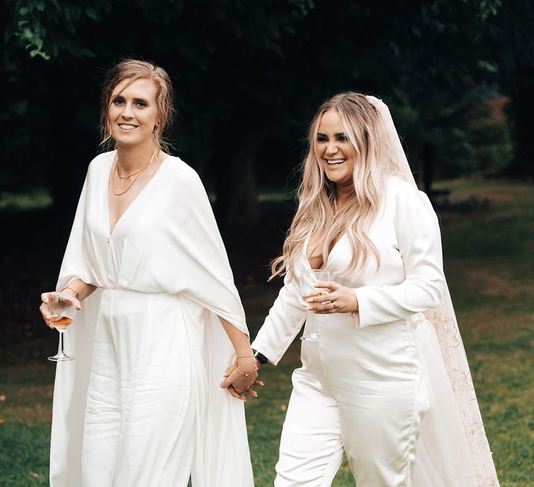 Brides walk along grass in bridal jumpsuits on their wedding day