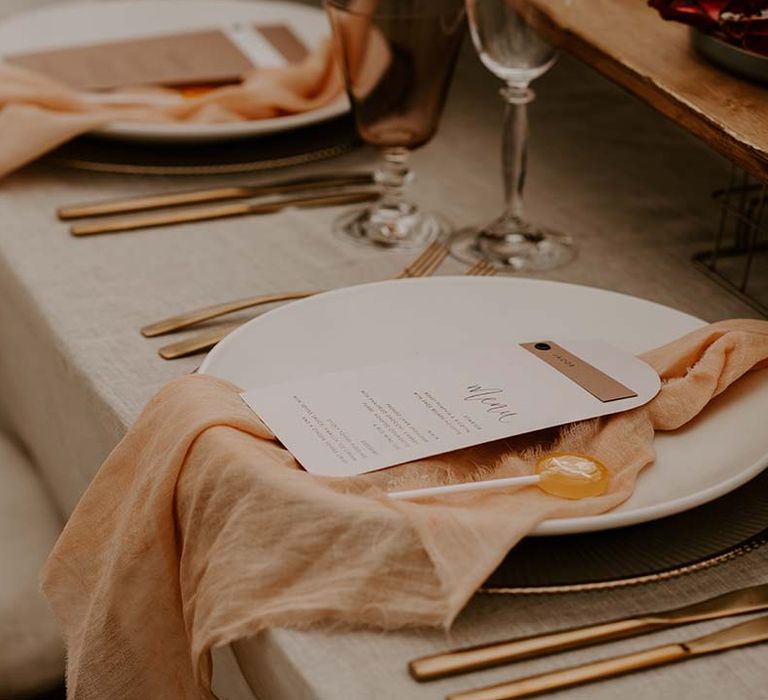 Elegant place setting with white tableware, natural linen napkin, brass cutlery, lollipop wedding favour and on the day stationery menu card