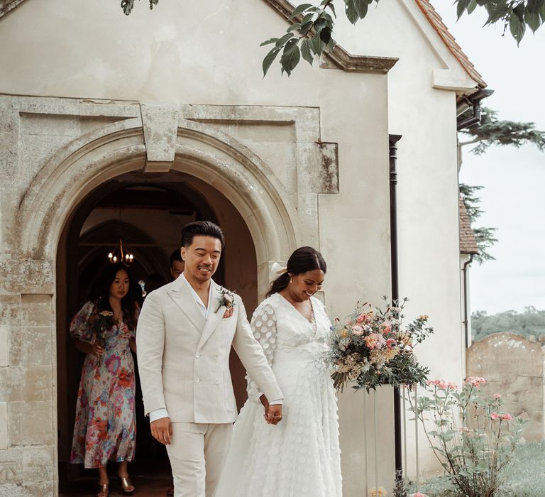 Groom in double breasted linen suit and white shawl collared shirt walks out of church holding hands long sleeve mesh spotted Vagabond wedding dress holding mixed bridal bouquet at Wasing Park wedding