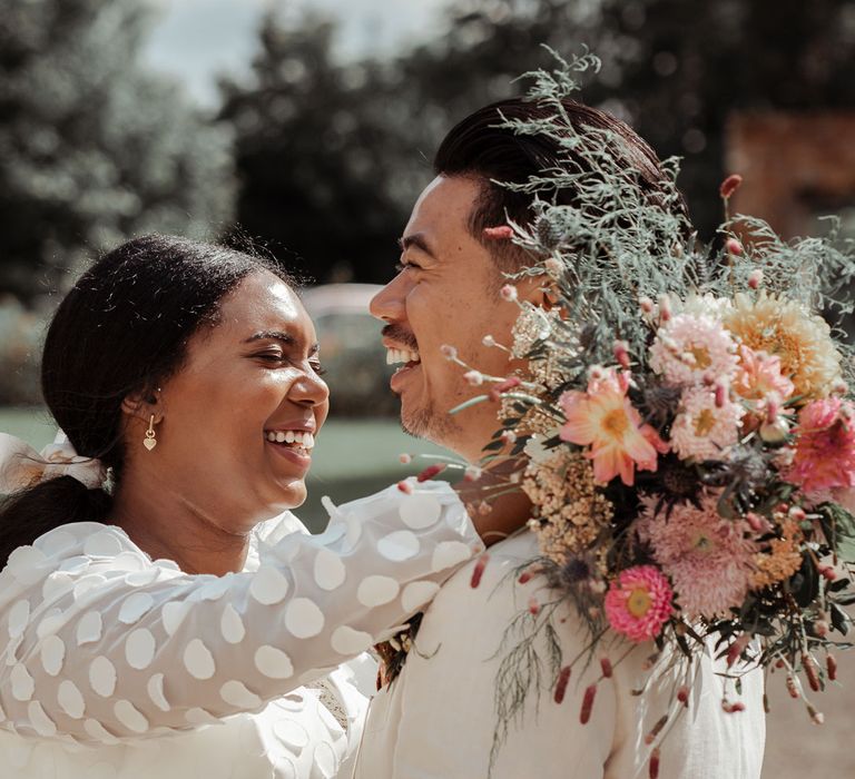 Bride in white long mesh sleeve spotted Vagabond wedding dress and bridal hair bow holding mixed bridal bouquet puts arms around neck of groom in linen suit as they both laugh standing outside at Wasing Park wedding