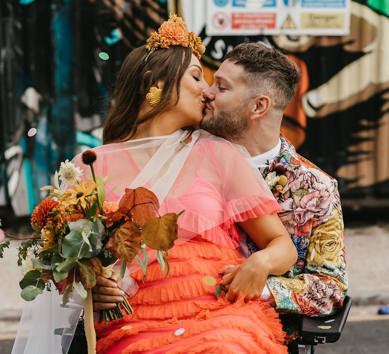 Bride in a coral wedding dress and cape veil kissing her C5/C6 Tetraplegic disabled groom in a wheelchair in Shoreditch 