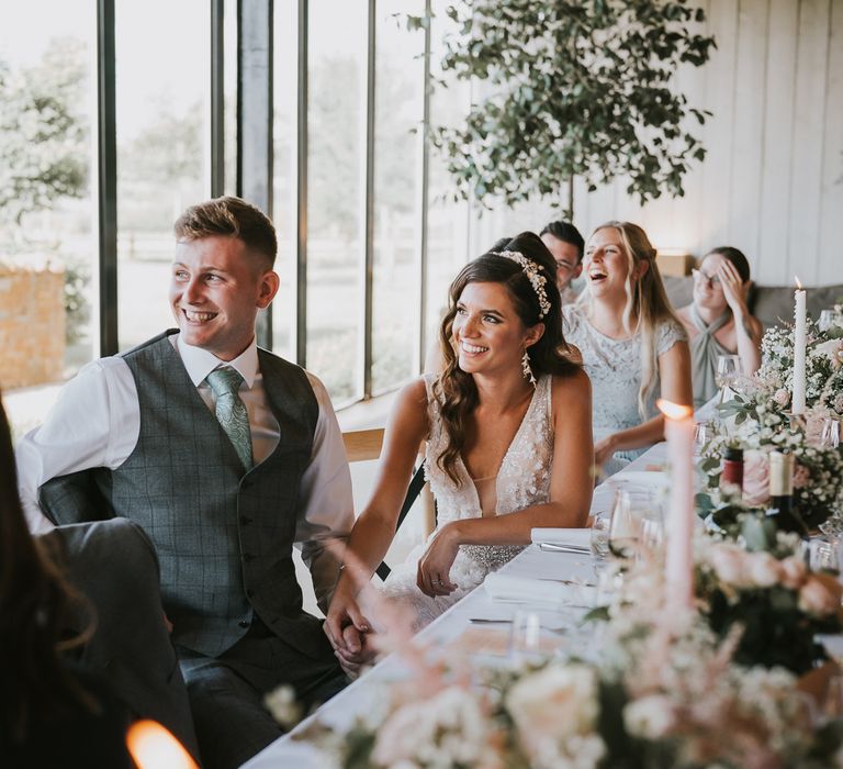 Groom in white shirt, grey waistcoat and mint green tie sits holding hands with bride in applique Berta wedding dress and headband during wedding breakfast at Primrose Hill Farm wedding