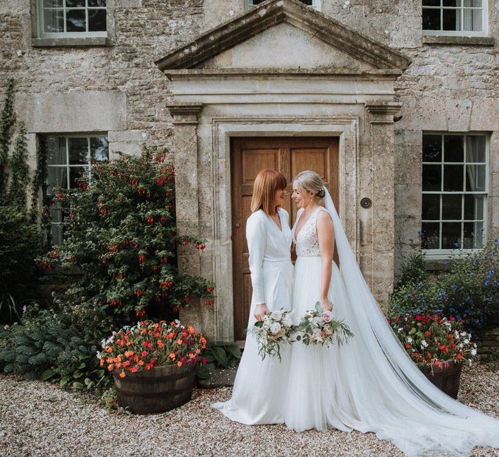 Two brides stand face to face, both holding bouquets outside stone house