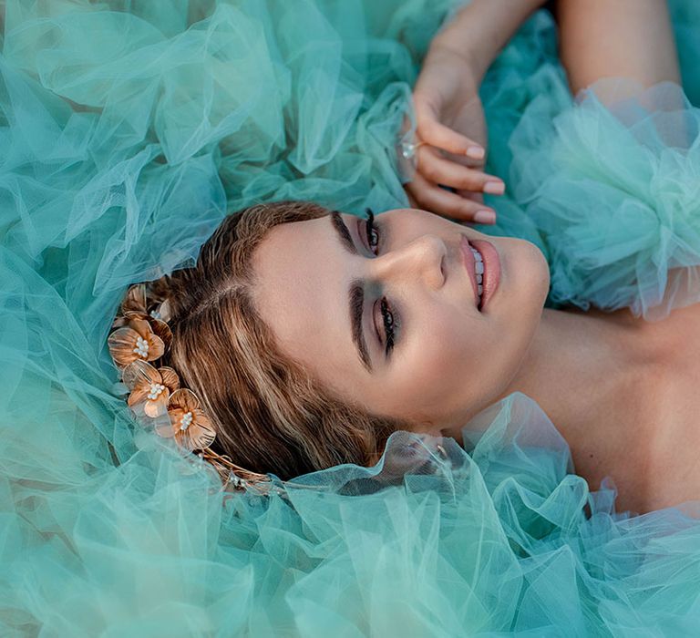 Bride with smokey brown and green eyeshadow and gold headdress laying on top of her mint green layered tulle wedding dress 