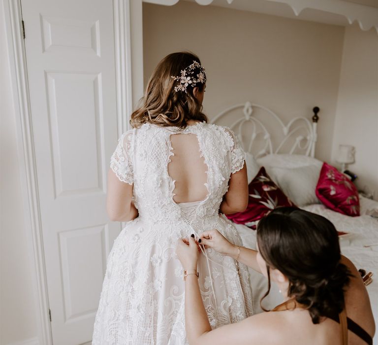 Bride in homemade lace wedding dress with open back has the back fastened by bridesmaid in rust cross back satin bridesmaid dress before DIY garden wedding in Bedfordshire