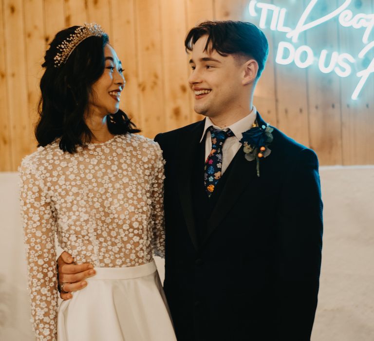 Bride in long sleeve applique wedding dress and headpiece stands with groom in dark blue suit, floral tie and floral buttonhole stand by neon sign at rustic wedding breakfast 