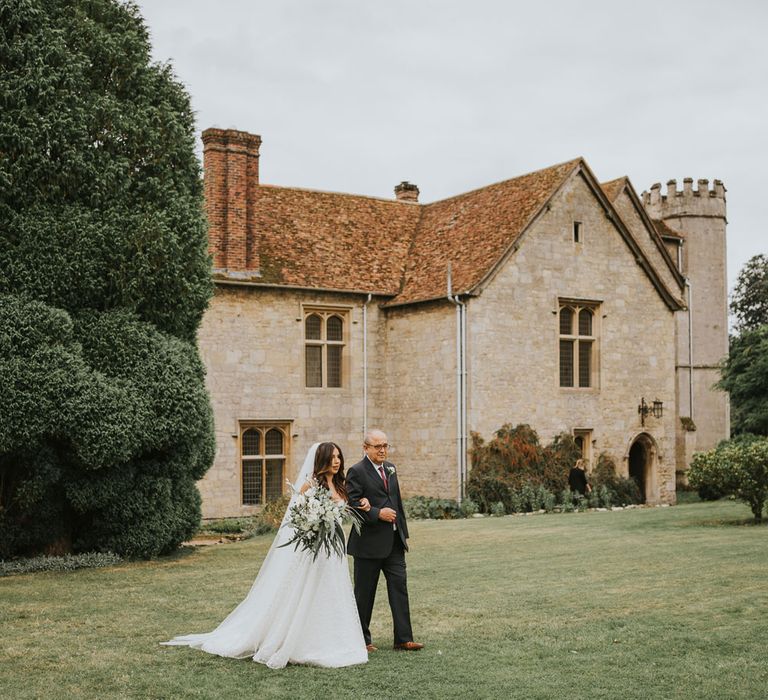 Bride in Pronovias wedding dress, Jimmy Choo wedding shoes and veil carrying white rose and green bridal bouquet walks arm-in-arm with man in dark suit and white rose buttonhole towards the aisle at outdoor wedding ceremony