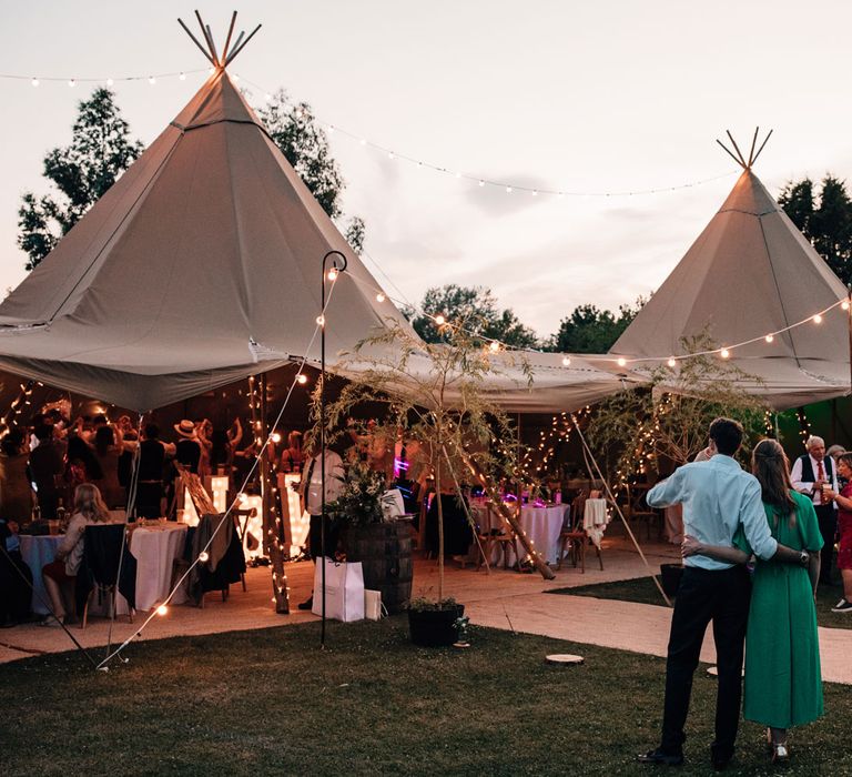 Wedding guests watch on as guests dance in fairy lit tipi for festival style wedding reception during golden hour